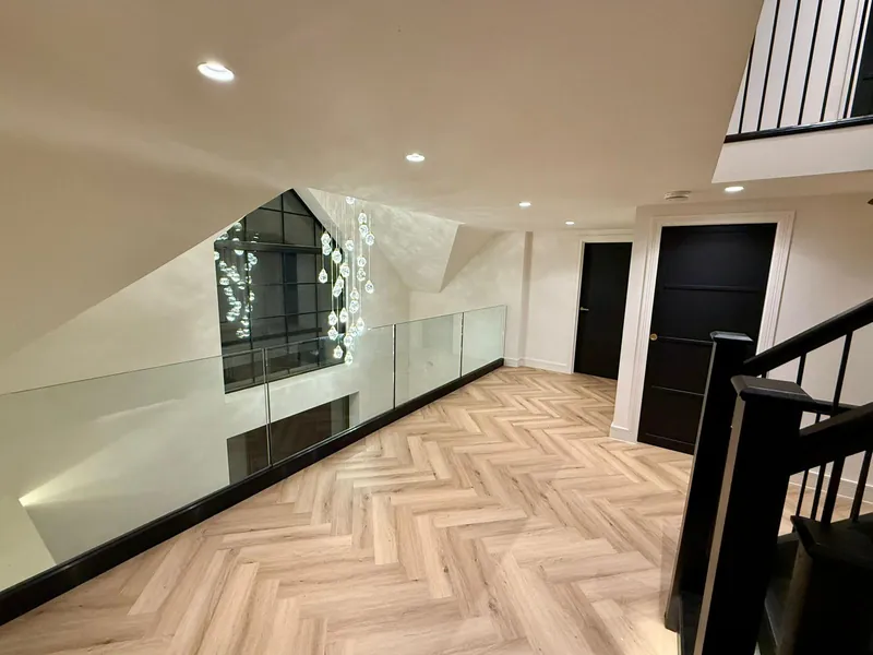 First-floor landing with a frameless glass balustrade, herringbone floor and a chandelier hanging in front of a tall glazed gable window