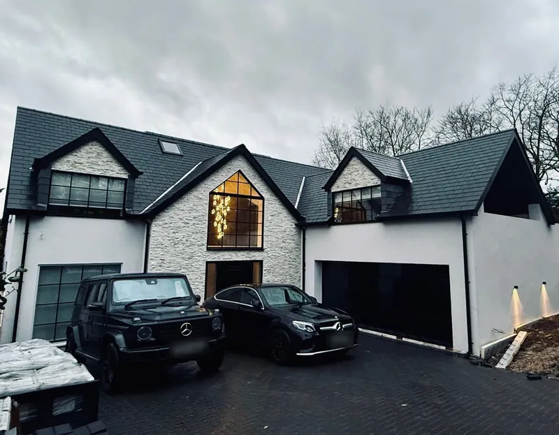 Front of a newly built house with a stone-clad gable, a tall black-framed window, a slate roof with dormers and a double garage