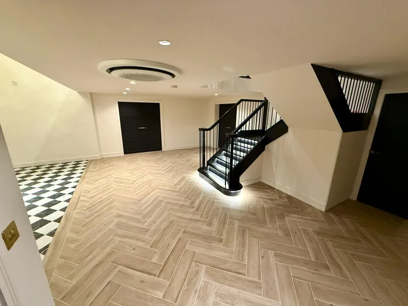 Large hall with a herringbone floor, a black staircase with lit treads, black double doors and a chequer-tiled area to one side